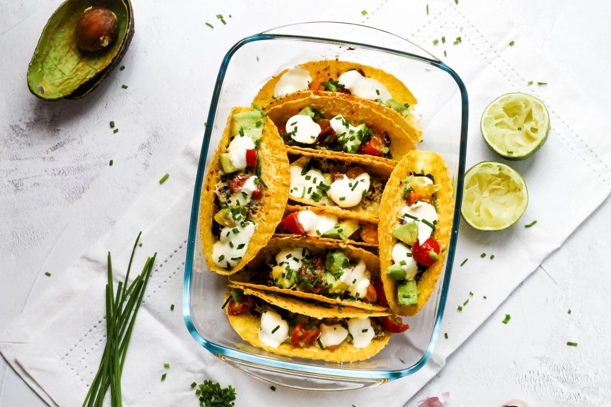 Landscape view of a tray of oven baked tacos with beans and cheese and a variety of topping, with herbs, limes and avocado skins to the side