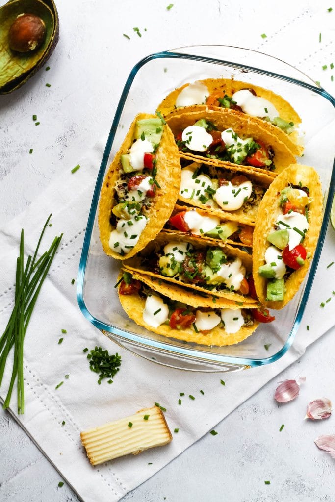 A top down image of baked tacos with avocado and herbs scattered around