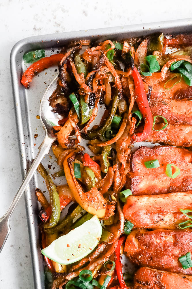 A top down view of a sheetpan with cooked roasted halloumi fajitas, highlighting the peppers and onions being spooned out